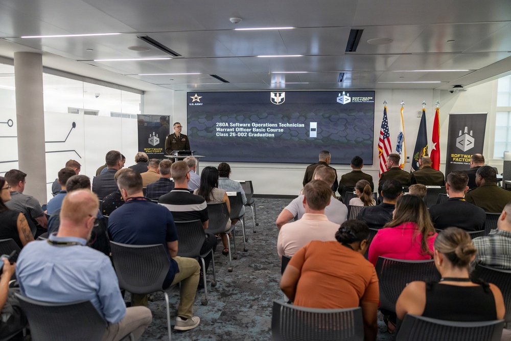 A military graduation ceremony is taking place at the U.S. Army Software Factory for the 280A Software Operations Technician Warrant Officer Basic Course, Class 26-002. A uniformed military officer is speaking at a podium in front of a large display screen showing the event details. An audience of approximately 30-40 attendees, including military personnel and civilians, are seated facing the speaker.