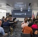 A military graduation ceremony is taking place at the U.S. Army Software Factory for the 280A Software Operations Technician Warrant Officer Basic Course, Class 26-002. A uniformed military officer is speaking at a podium in front of a large display screen showing the event details. An audience of approximately 30-40 attendees, including military personnel and civilians, are seated facing the speaker.