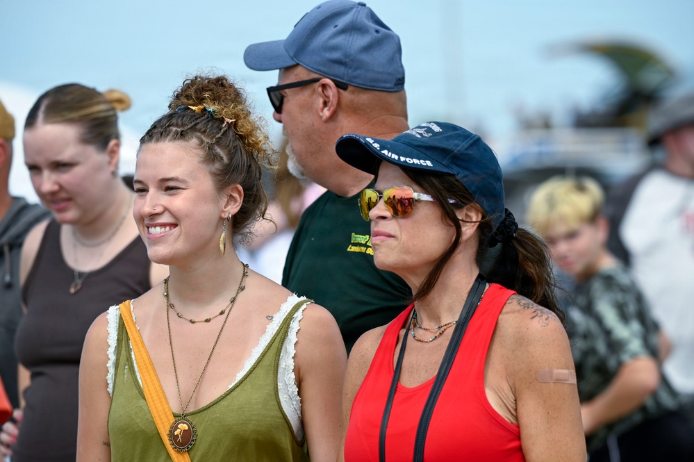 A group of people gathered at what appears to be an outdoor public event or festival. Two women are prominently featured in the foreground, one smiling broadly wearing a green tank top with layered necklaces, and another wearing a red tank top, sunglasses, and a U.S. Air Force cap. Additional attendees are visible in the background, slightly out of focus.