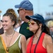A group of people gathered at what appears to be an outdoor public event or festival. Two women are prominently featured in the foreground, one smiling broadly wearing a green tank top with layered necklaces, and another wearing a red tank top, sunglasses, and a U.S. Air Force cap. Additional attendees are visible in the background, slightly out of focus.
