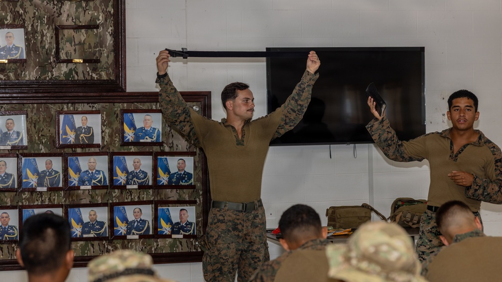 A military training or instructional session is taking place indoors, where a service member in MARPAT digital camouflage uniform is demonstrating a technique by holding a black strap or belt overhead with both arms raised. A second service member stands nearby observing or assisting, while an audience of seated personnel in mixed camouflage uniforms watches. The room is decorated with a wall display of framed official military portraits and photographs.