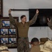 A military training or instructional session is taking place indoors, where a service member in MARPAT digital camouflage uniform is demonstrating a technique by holding a black strap or belt overhead with both arms raised. A second service member stands nearby observing or assisting, while an audience of seated personnel in mixed camouflage uniforms watches. The room is decorated with a wall display of framed official military portraits and photographs.