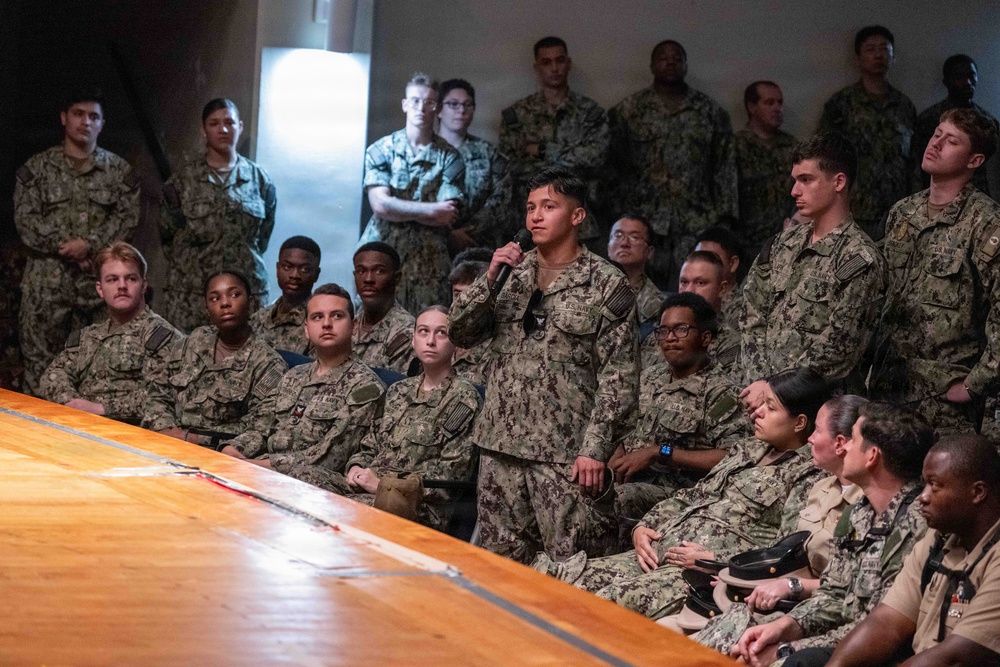 A large group of U.S. Navy personnel in Navy Working Uniform (NWU) Type III camouflage are gathered in what appears to be a briefing or all-hands meeting. One service member standing in the center is speaking into a handheld microphone, suggesting a question-and-answer session or formal address. Senior personnel in khaki uniforms are visible seated in the foreground near a wooden table.