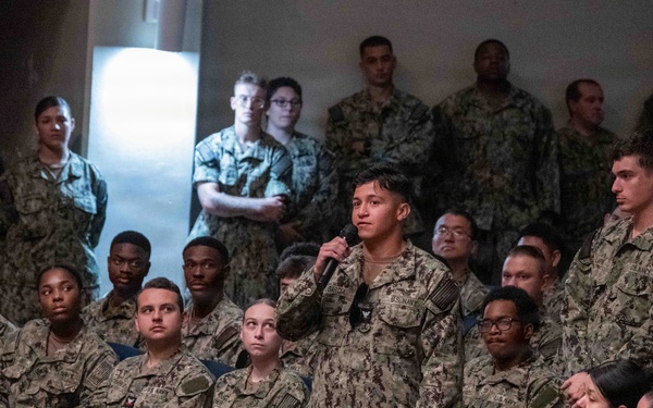 A large group of U.S. Navy personnel in Navy Working Uniform (NWU) Type III camouflage are gathered in what appears to be a briefing or all-hands meeting. One service member standing in the center is speaking into a handheld microphone, suggesting a question-and-answer session or formal address. Senior personnel in khaki uniforms are visible seated in the foreground near a wooden table.