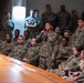 A large group of U.S. Navy personnel in Navy Working Uniform (NWU) Type III camouflage are gathered in what appears to be a briefing or all-hands meeting. One service member standing in the center is speaking into a handheld microphone, suggesting a question-and-answer session or formal address. Senior personnel in khaki uniforms are visible seated in the foreground near a wooden table.