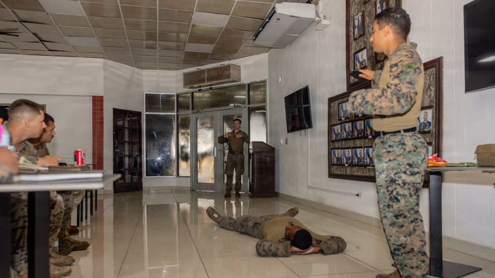 Military personnel, appearing to be U.S. Marines in digital camouflage uniforms, are engaged in what appears to be a training or demonstration exercise inside a formal building. One individual is lying prone on the floor as part of the demonstration, while an instructor stands near the entrance gesturing, and others observe. The setting appears to be a military headquarters or administrative building, evidenced by framed portraits and plaques on the wall.