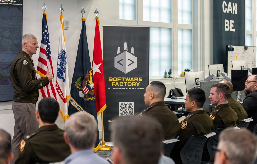 A military officer in U.S. Army uniform is presenting or briefing a group of uniformed soldiers and civilians at what appears to be the Army Software Factory facility. The setting is a modern office/tech environment with multiple flags displayed including the U.S. flag and Army-related flags. The banner behind the speaker reads 'Software Factory - By Soldiers. For Soldiers.'