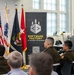 A military officer in U.S. Army uniform is presenting or briefing a group of uniformed soldiers and civilians at what appears to be the Army Software Factory facility. The setting is a modern office/tech environment with multiple flags displayed including the U.S. flag and Army-related flags. The banner behind the speaker reads 'Software Factory - By Soldiers. For Soldiers.'