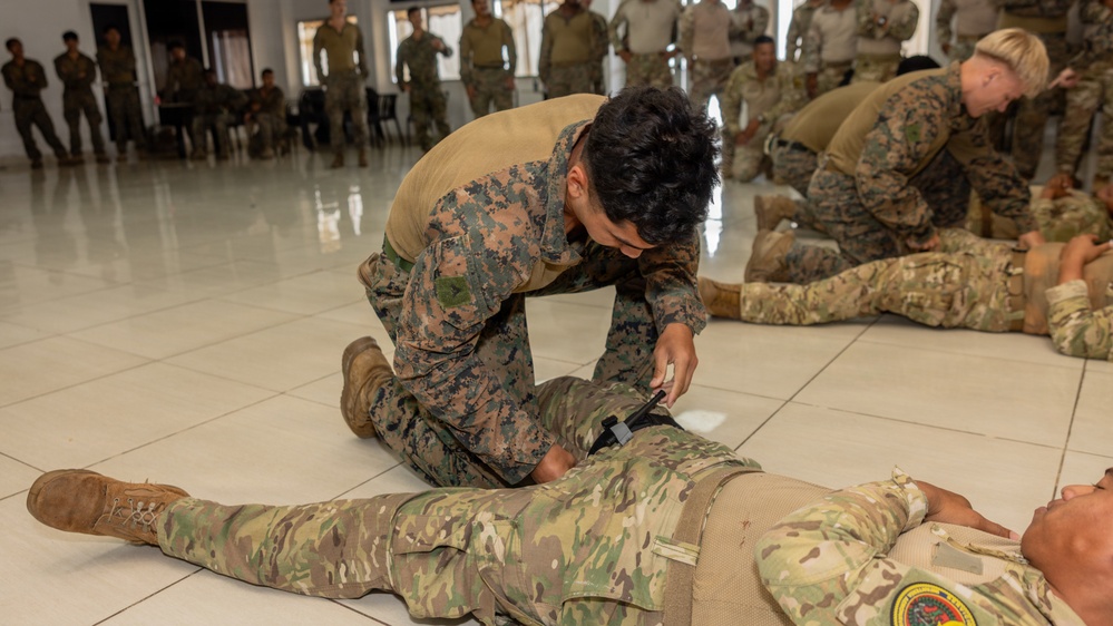 Military personnel are conducting a combat casualty care or tactical combat medicine training exercise in an indoor facility. A soldier in digital camouflage is kneeling over a simulated casualty, applying what appears to be a tourniquet to the leg. Multiple pairs of soldiers are practicing the same procedure throughout the room, with additional personnel observing from the background.