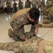 Military personnel are conducting a combat casualty care or tactical combat medicine training exercise in an indoor facility. A soldier in digital camouflage is kneeling over a simulated casualty, applying what appears to be a tourniquet to the leg. Multiple pairs of soldiers are practicing the same procedure throughout the room, with additional personnel observing from the background.