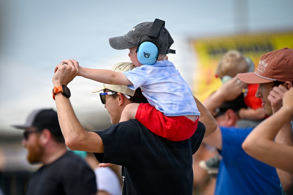 A young child wearing blue ear protection headphones and a gray baseball cap sits on an adult's shoulders at what appears to be an outdoor airshow or loud public event. The child is holding the adult's hands for balance while looking forward. Multiple spectators are visible in the background, some also shielding their eyes or wearing ear protection.