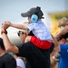 A young child wearing blue ear protection headphones and a gray baseball cap sits on an adult's shoulders at what appears to be an outdoor airshow or loud public event. The child is holding the adult's hands for balance while looking forward. Multiple spectators are visible in the background, some also shielding their eyes or wearing ear protection.