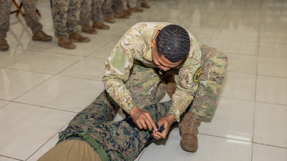 A soldier in MultiCam/OCP camouflage uniform is kneeling on a tiled floor, applying what appears to be a tourniquet or medical device to the leg of a person lying prone in MARPAT (Marine) camouflage. The setting appears to be an indoor training environment, with a line of uniformed personnel visible in the background observing the demonstration. This appears to be a tactical combat casualty care (TCCC) or first aid training exercise.
