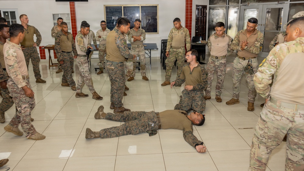 Military personnel in various camouflage uniforms are gathered in an indoor facility conducting what appears to be a combat training or tactical demonstration session. One soldier is lying on the floor while another kneels beside them, suggesting a scenario involving casualty care, combatives, or detainee handling instruction. A group of approximately 15-20 service members surrounds the demonstration, observing the technique being taught.