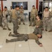 Military personnel in various camouflage uniforms are gathered in an indoor facility conducting what appears to be a combat training or tactical demonstration session. One soldier is lying on the floor while another kneels beside them, suggesting a scenario involving casualty care, combatives, or detainee handling instruction. A group of approximately 15-20 service members surrounds the demonstration, observing the technique being taught.
