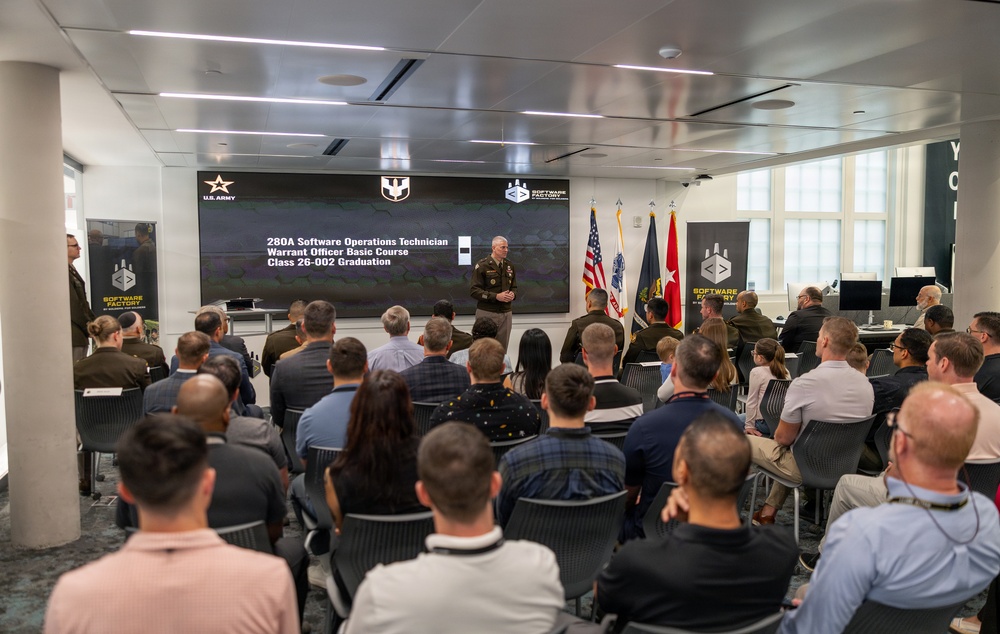 A U.S. Army graduation ceremony for the 280A Software Operations Technician Warrant Officer Basic Course, Class 26-002, is taking place at the Army Software Factory. A uniformed officer stands at the front of the room addressing an audience of approximately 40-50 attendees seated in rows. The event is held in a modern facility with branded 'Software Factory' signage and military flags displayed prominently.