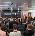 A U.S. Army graduation ceremony for the 280A Software Operations Technician Warrant Officer Basic Course, Class 26-002, is taking place at the Army Software Factory. A uniformed officer stands at the front of the room addressing an audience of approximately 40-50 attendees seated in rows. The event is held in a modern facility with branded 'Software Factory' signage and military flags displayed prominently.