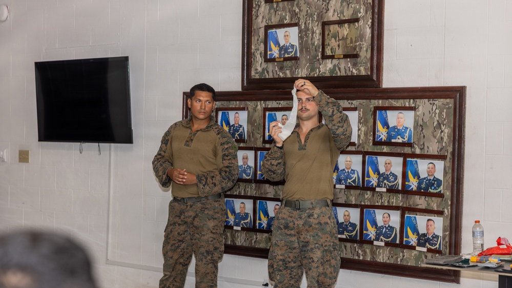 Two military personnel in camouflage uniforms are standing in front of a wall displaying framed official portraits of military officers. One individual is holding up what appears to be a white object or bandage, suggesting a demonstration or training session. The setting appears to be an indoor military facility or briefing room.
