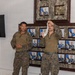 Two military personnel in camouflage uniforms are standing in front of a wall displaying framed official portraits of military officers. One individual is holding up what appears to be a white object or bandage, suggesting a demonstration or training session. The setting appears to be an indoor military facility or briefing room.