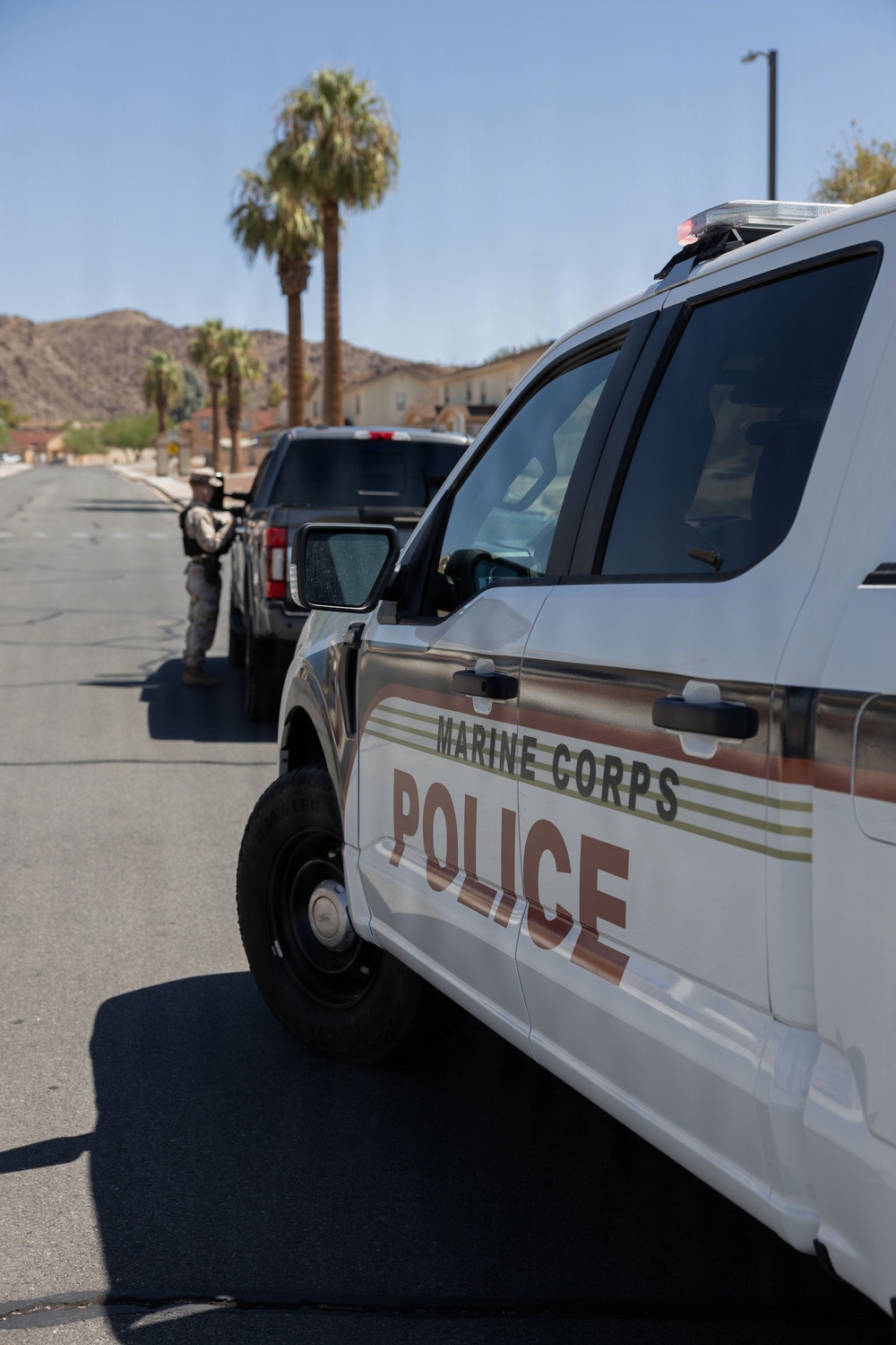 A Marine Corps Police vehicle is parked on a street within what appears to be a military base installation, identifiable by the desert landscape and palm trees in the background. A uniformed personnel member is visible in the background standing near a dark SUV, appearing to conduct a traffic stop or checkpoint. The setting suggests a residential or administrative area of a Marine Corps base in a desert region.
