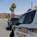 A Marine Corps Police vehicle is parked on a street within what appears to be a military base installation, identifiable by the desert landscape and palm trees in the background. A uniformed personnel member is visible in the background standing near a dark SUV, appearing to conduct a traffic stop or checkpoint. The setting suggests a residential or administrative area of a Marine Corps base in a desert region.