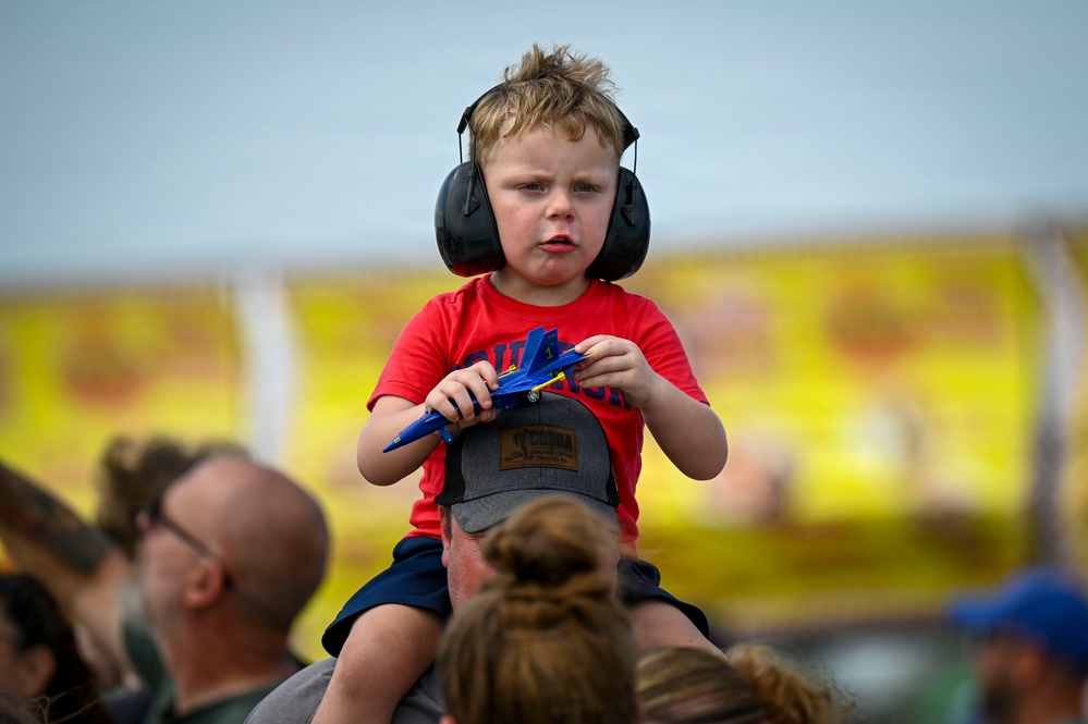 A young boy, approximately 3-5 years old, sits on an adult's shoulders at what appears to be an air show. He is wearing black ear protection headphones and a red t-shirt, holding a blue toy jet aircraft that resembles a Blue Angels F/A-18 model. The background shows a crowd of spectators and yellow signage consistent with an outdoor public event.