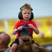 A young boy, approximately 3-5 years old, sits on an adult's shoulders at what appears to be an air show. He is wearing black ear protection headphones and a red t-shirt, holding a blue toy jet aircraft that resembles a Blue Angels F/A-18 model. The background shows a crowd of spectators and yellow signage consistent with an outdoor public event.