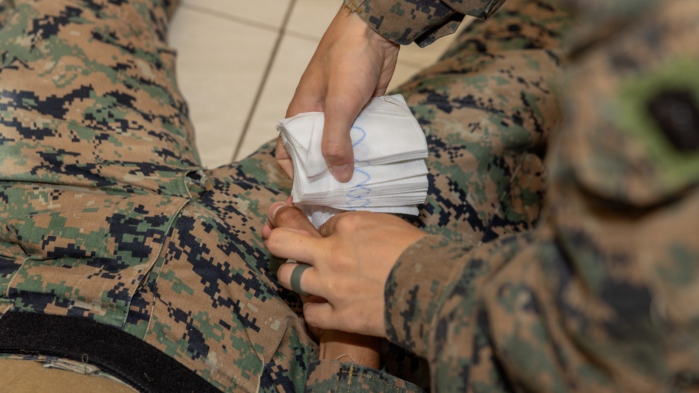 The image shows what appears to be a military medical training or first aid scenario involving two individuals in U.S. Marine Corps MARPAT (digital camouflage) uniforms. One person is applying what appears to be gauze or wound packing material to another person's torso area. This is consistent with tactical combat casualty care (TCCC) training or a medical aid drill.