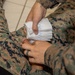 The image shows what appears to be a military medical training or first aid scenario involving two individuals in U.S. Marine Corps MARPAT (digital camouflage) uniforms. One person is applying what appears to be gauze or wound packing material to another person's torso area. This is consistent with tactical combat casualty care (TCCC) training or a medical aid drill.