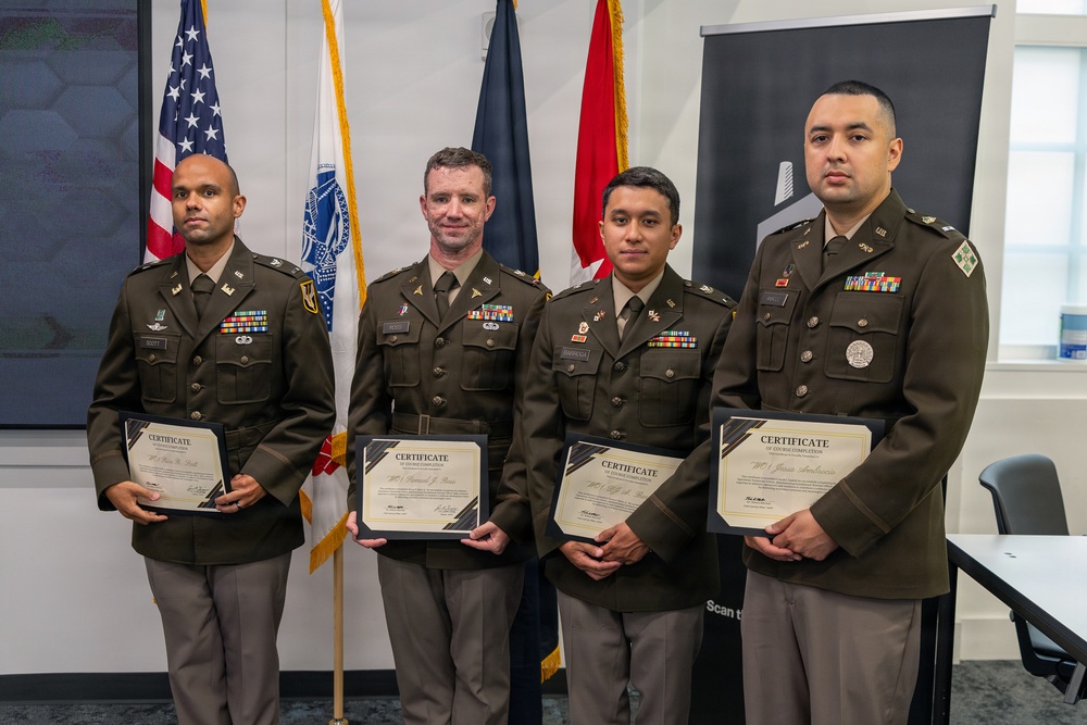 Four U.S. Army soldiers in Army Service Uniforms (ASUs) pose together holding certificates of course completion. They are standing in front of multiple flags including the U.S. flag, a military branch flag, and what appears to be a red flag, likely in an official military facility. The occasion appears to be a formal graduation or course completion ceremony.