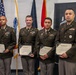 Four U.S. Army soldiers in Army Service Uniforms (ASUs) pose together holding certificates of course completion. They are standing in front of multiple flags including the U.S. flag, a military branch flag, and what appears to be a red flag, likely in an official military facility. The occasion appears to be a formal graduation or course completion ceremony.