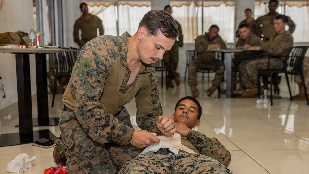 A military training exercise is taking place in an indoor facility where one service member in MARPAT camouflage uniform is practicing first aid or combat casualty care on another service member lying on the floor. The standing individual appears to be applying a bandage or dressing to the neck/shoulder area of the person on the ground. Additional service members are visible in the background, seated at tables and observing or participating in similar training activities.