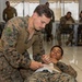 A military training exercise is taking place in an indoor facility where one service member in MARPAT camouflage uniform is practicing first aid or combat casualty care on another service member lying on the floor. The standing individual appears to be applying a bandage or dressing to the neck/shoulder area of the person on the ground. Additional service members are visible in the background, seated at tables and observing or participating in similar training activities.
