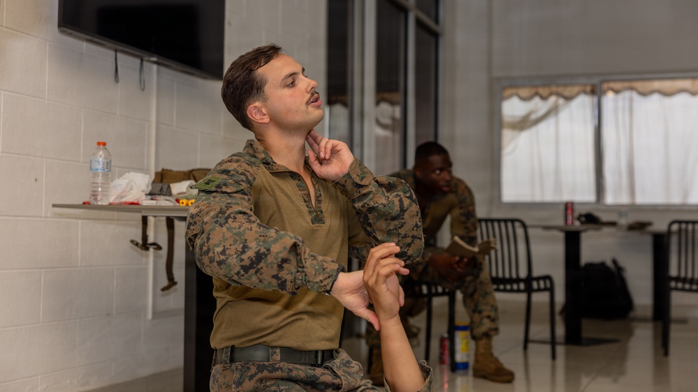 A U.S. Marine in MARPAT camouflage uniform is seated in what appears to be an indoor break or waiting area, with his hand raised to his chin in a thoughtful or attentive pose. A second Marine is visible in the background, seated and appearing to read or review materials. The setting suggests a briefing room, rest area, or administrative space at a military installation.