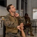 A U.S. Marine in MARPAT camouflage uniform is seated in what appears to be an indoor break or waiting area, with his hand raised to his chin in a thoughtful or attentive pose. A second Marine is visible in the background, seated and appearing to read or review materials. The setting suggests a briefing room, rest area, or administrative space at a military installation.