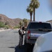 A military personnel member in camouflage uniform is standing beside a gray pickup truck on a residential street in what appears to be a desert Southwest location. The soldier is carrying equipment on their back and appears to be interacting with the vehicle's driver-side window. The setting features palm trees, rocky desert hills, and clear blue skies consistent with Arizona or Southern California.