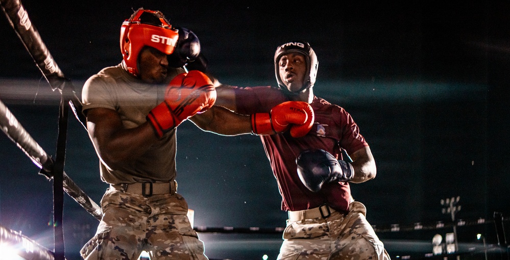 Two boxers are competing in a boxing match inside a ring under dramatic lighting against a dark background. Both fighters are wearing military-style camouflage or uniform pants alongside boxing gear, suggesting this may be a military boxing event. One fighter is landing or throwing a punch while the other reacts to the impact.