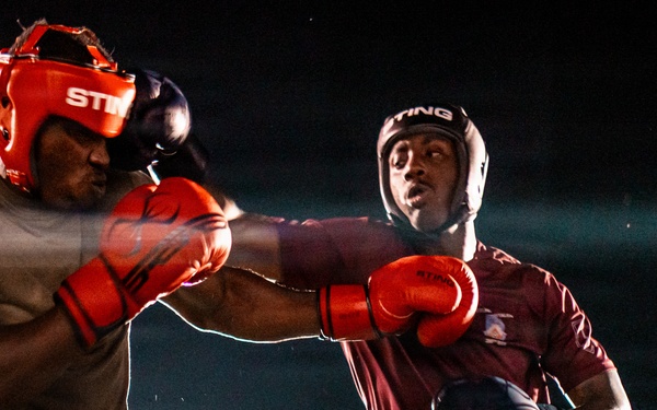 Two boxers are competing in a boxing match inside a ring under dramatic lighting against a dark background. Both fighters are wearing military-style camouflage or uniform pants alongside boxing gear, suggesting this may be a military boxing event. One fighter is landing or throwing a punch while the other reacts to the impact.