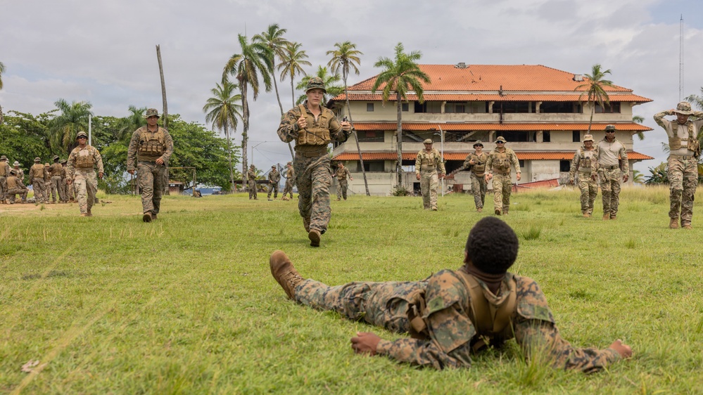 Military personnel in camouflage uniforms are conducting outdoor training exercises on a grassy field near a multi-story building with a red-tiled roof and palm trees in the background. One soldier is prone on the ground in the foreground while others are walking, jogging, and standing across the field. The setting appears to be a tropical military installation.