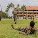 Military personnel in camouflage uniforms are conducting outdoor training exercises on a grassy field near a multi-story building with a red-tiled roof and palm trees in the background. One soldier is prone on the ground in the foreground while others are walking, jogging, and standing across the field. The setting appears to be a tropical military installation.