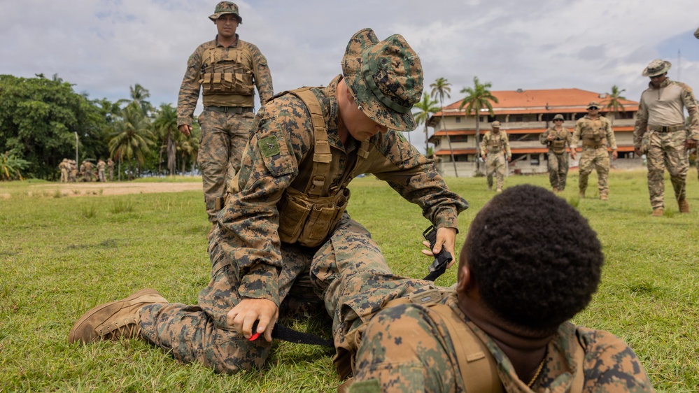Military personnel in camouflage uniforms are conducting what appears to be a combat casualty care or tactical first aid training exercise on a grass field. One soldier kneels over another who is lying on the ground, appearing to apply or demonstrate tourniquet application. Additional uniformed personnel observe in the background near a building surrounded by tropical vegetation.