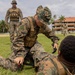 Military personnel in camouflage uniforms are conducting what appears to be a combat casualty care or tactical first aid training exercise on a grass field. One soldier kneels over another who is lying on the ground, appearing to apply or demonstrate tourniquet application. Additional uniformed personnel observe in the background near a building surrounded by tropical vegetation.