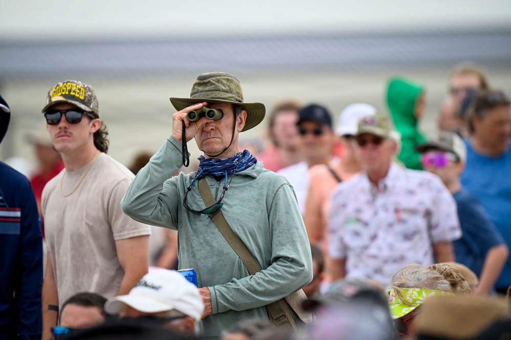 A crowd of spectators gathered at what appears to be an outdoor event, likely an airshow or similar large public gathering near a tarmac or open field. The central figure is a middle-aged person using binoculars to observe something in the distance, wearing sun-protective clothing including a wide-brim hat and neck gaiter. The crowd is diverse in age and dressed in casual warm-weather attire.