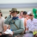 A crowd of spectators gathered at what appears to be an outdoor event, likely an airshow or similar large public gathering near a tarmac or open field. The central figure is a middle-aged person using binoculars to observe something in the distance, wearing sun-protective clothing including a wide-brim hat and neck gaiter. The crowd is diverse in age and dressed in casual warm-weather attire.