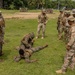 Military personnel from what appear to be two different forces — U.S. Marines in MARPAT camouflage and soldiers in a tan/multicam pattern — are conducting a joint ground fighting or combatives training exercise on a grass field. Two participants are on the ground demonstrating a grappling or restraint technique while others form a circle observing. The tropical environment, palm trees, and building architecture suggest a location in Central America or the Caribbean.