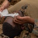 The image shows military personnel in camouflage uniforms administering first aid to a fellow service member lying on the ground. Two individuals are applying what appears to be a white gauze bandage to the face/eye area of the person on the ground. This appears to be either a combat casualty care scenario or a tactical combat casualty care (TCCC) training exercise.
