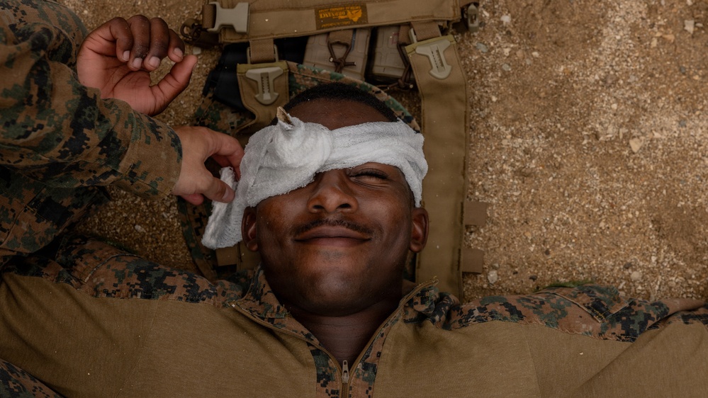 A military service member in MARPAT digital camouflage uniform is lying on the ground receiving first aid treatment to his eye area. Another service member is applying or adjusting a white gauze bandage/dressing over the casualty's eye. The expression on the casualty's face appears relaxed or slightly smiling, suggesting this may be a training exercise rather than an actual combat casualty.