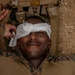 A military service member in MARPAT digital camouflage uniform is lying on the ground receiving first aid treatment to his eye area. Another service member is applying or adjusting a white gauze bandage/dressing over the casualty's eye. The expression on the casualty's face appears relaxed or slightly smiling, suggesting this may be a training exercise rather than an actual combat casualty.