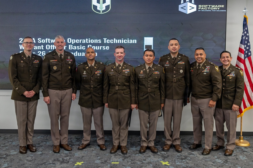 A group of eight U.S. Army personnel in dress green uniforms pose for a formal group photo at a graduation ceremony. The event is identified by the presentation screen behind them as a '25A Software Operations Technician Warrant Officer Basic Course' graduation. An American flag is visible on the right side of the frame, and the Army Software Factory logo is displayed on the screen.