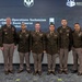 A group of eight U.S. Army personnel in dress green uniforms pose for a formal group photo at a graduation ceremony. The event is identified by the presentation screen behind them as a '25A Software Operations Technician Warrant Officer Basic Course' graduation. An American flag is visible on the right side of the frame, and the Army Software Factory logo is displayed on the screen.